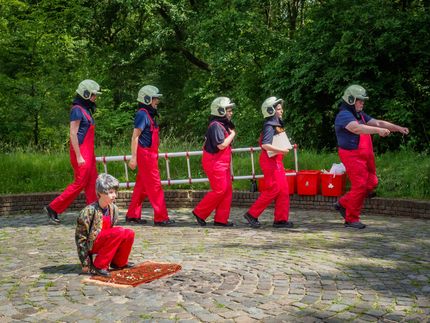 People in red pants and helmets walking in a park; one sits on a mat in the foreground.