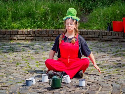 Person sitting cross-legged outdoors with a green hat and red overalls, surrounded by metal cups and kettles