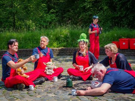 People in red overalls sit on a picnic blanket in a grassy park, eating and talking around cups and bags.