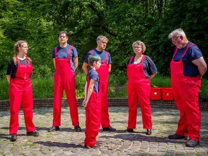 Six people in red overalls stand outdoors on a cobblestone path in front of trees.