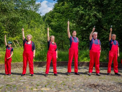 Six people in red overalls stand outdoors with one arm raised in a wooded park.