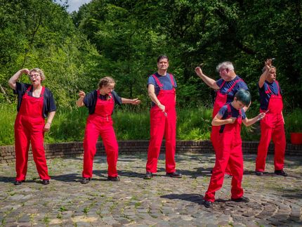 Six dancers in red overalls perform on a stone path in a green park.