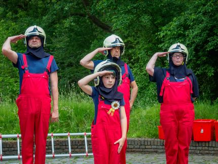 Four helmeted people in red overalls stand outdoors, saluting in front of green trees.