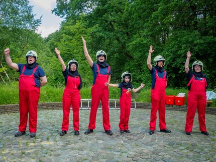 Six people in red outfits and helmets stand outdoors, smiling and waving on a cobblestone path.