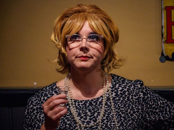Person with blond hair and glasses holding a drink indoors against a tan wall