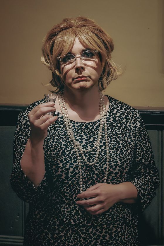 Person in patterned dress holding a spoon, standing against a wall with a neutral expression.