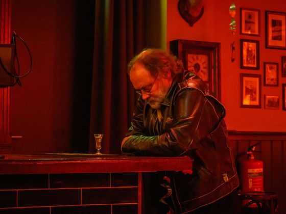 Man in a black leather jacket leaning on a bar in a red-lit room