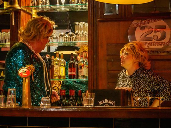 Bartender talking with a customer in a warmly lit bar, with shelves of bottles behind them.