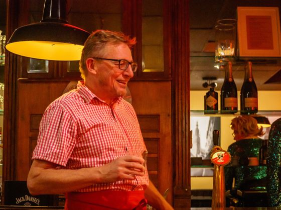 Man in red checkered shirt holding a drink at a warmly lit bar