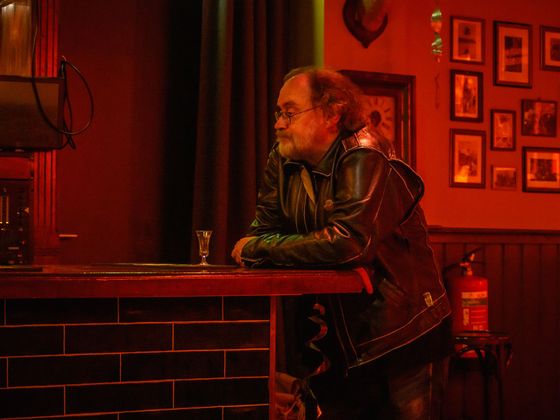 Man in a black leather jacket sitting at a red-lit bar, leaning on the counter, with framed photos on the wall.