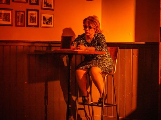 Woman seated on a bar stool at a small table in a warmly lit café or pub