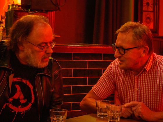 Two men talking at a table in a dim red-lit bar, with drinks and a brick wall behind them
