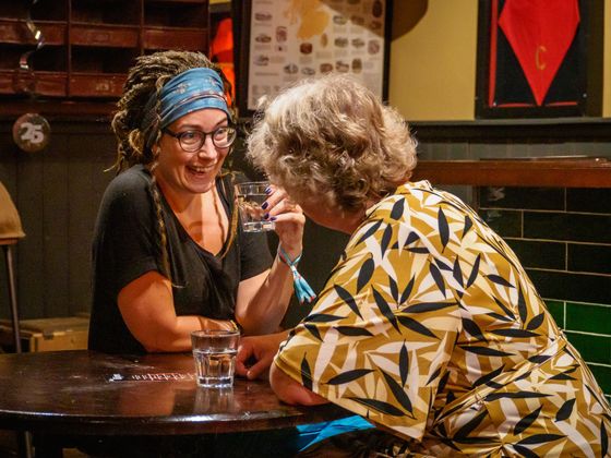Two people chatting at a bar, one wearing glasses and a blue headscarf, with drinks on the table.