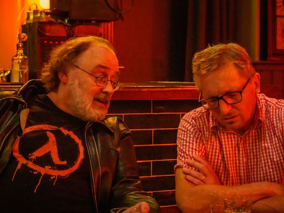 Two men talking at a table in a dim, red-lit setting; one wears a black jacket with an orange logo.