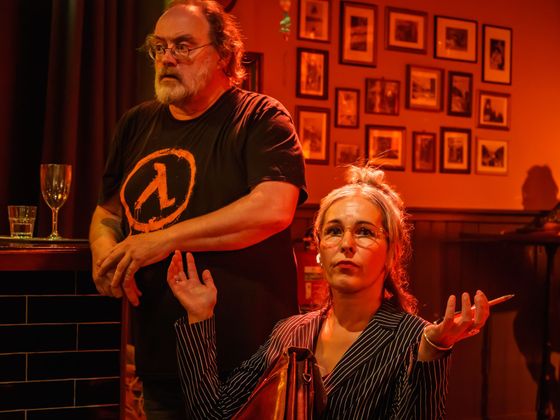 Two people in a dim, orange-lit room, one standing by a bar and one seated gesturing.