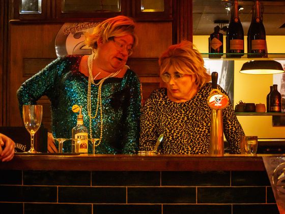Two people leaning at a bar, one in a teal sequined top and one in a patterned blouse, under warm lights.