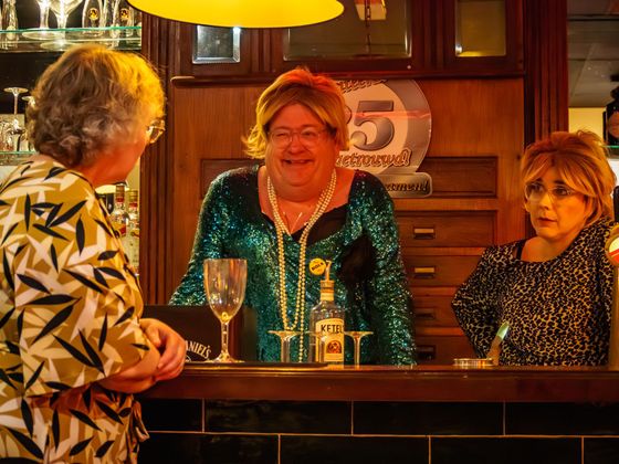 Three people chatting at a warmly lit bar, with drinks on the counter and a festive wooden interior.