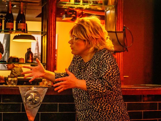 Person behind a bar gesturing while speaking, with bottles and warm red lighting in the background.