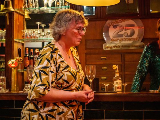 Woman in a patterned shirt stands at a bar with bottles, glasses, and warm amber lighting.