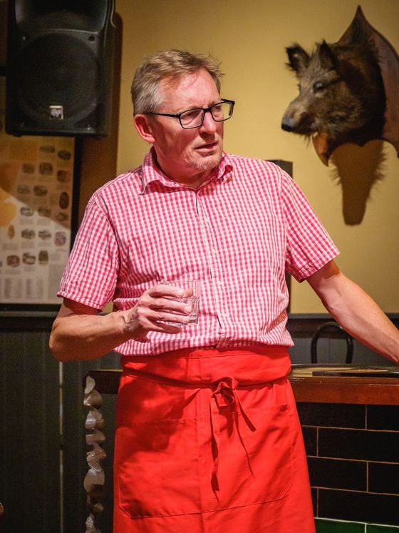 Man in red apron and checkered shirt speaking indoors beside a mounted boar head