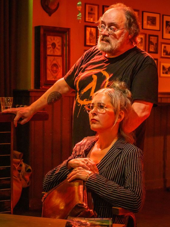 Two people seated indoors in warm orange light, looking off to the side in a cozy room with framed pictures.