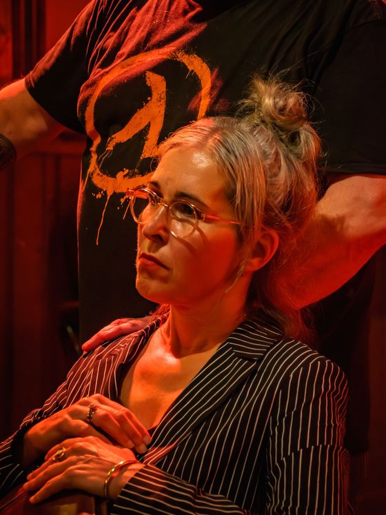 Woman with glasses seated under red stage lighting, hands clasped, looking to the side