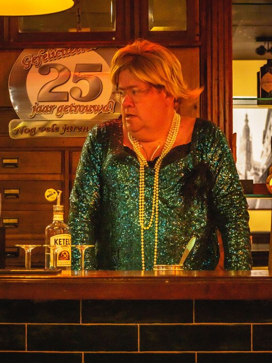 Woman in a green sequined jacket behind a bar with a bottle and a 25th anniversary sign behind her