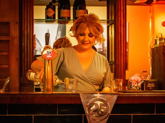 Bartender smiling behind a wooden bar, pouring a drink in a warmly lit pub.