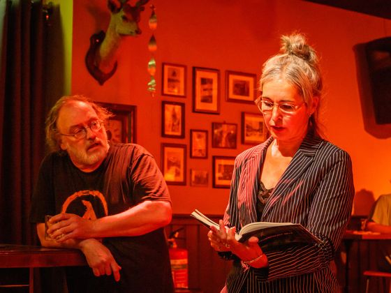 Two people in warm red lighting, one reading from a notebook beside a wall of framed pictures.