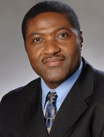Professional headshot of a man in a dark suit, blue shirt, and patterned tie against a gray background
