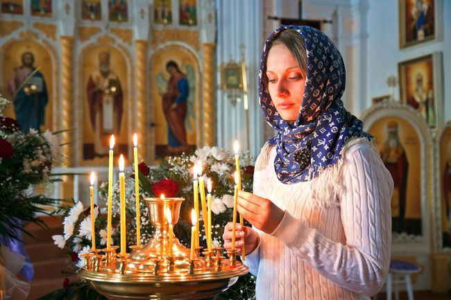 Woman in headscarf lighting candles in an Orthodox church, altar in background.