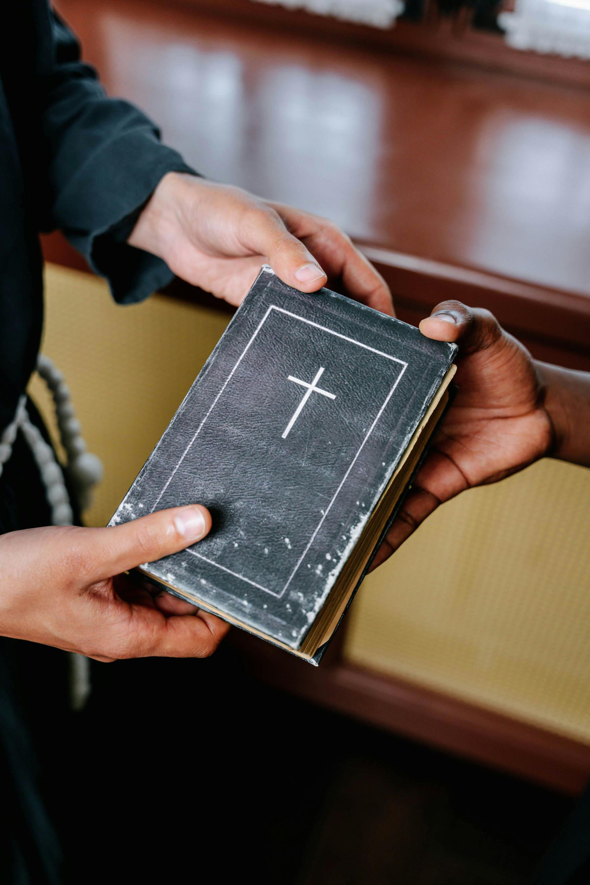 Hands passing a dark book with a white cross, possibly a Bible, indoors.