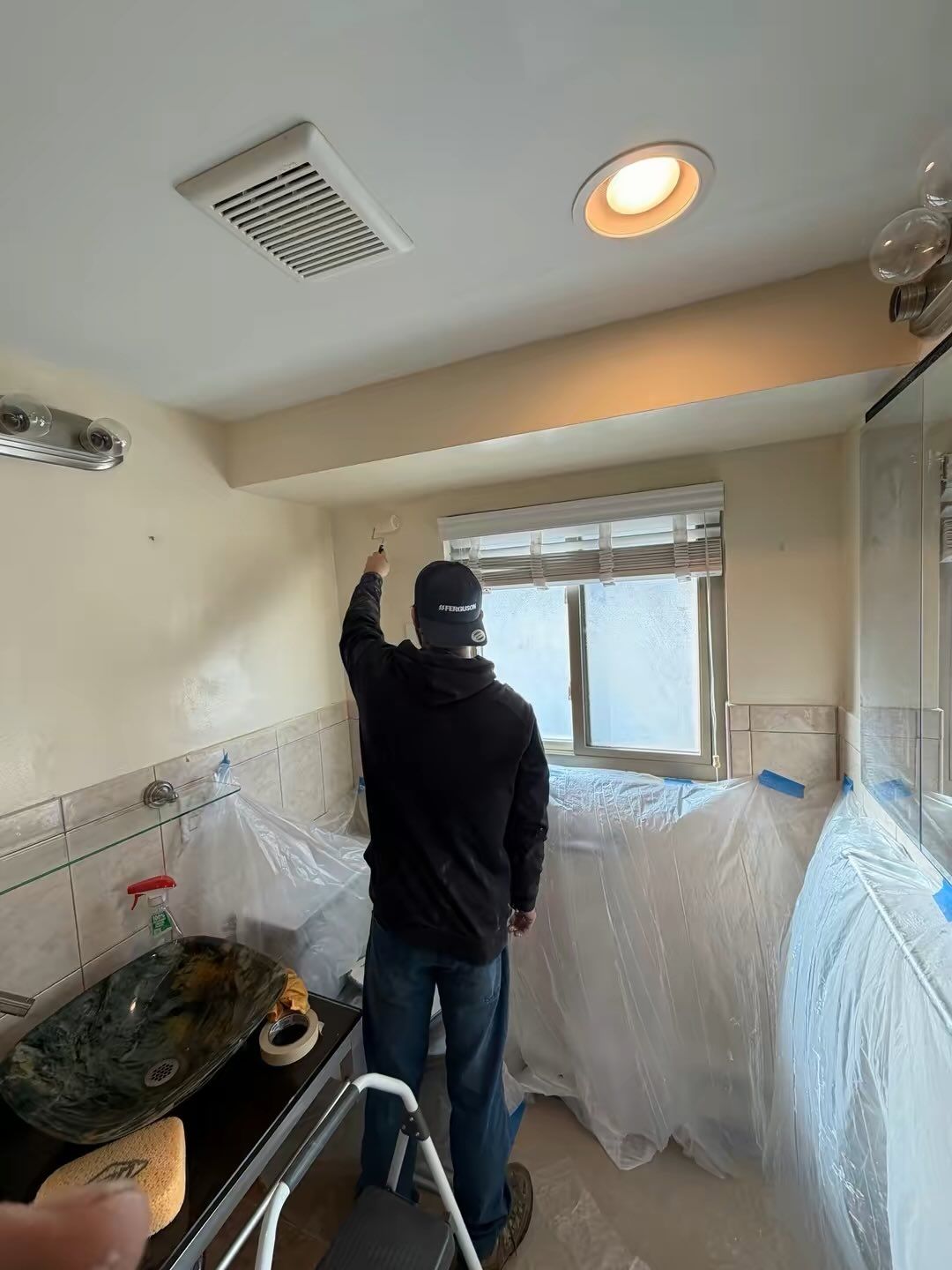Person painting a beige bathroom ceiling near a window, with plastic covering the sink and walls.