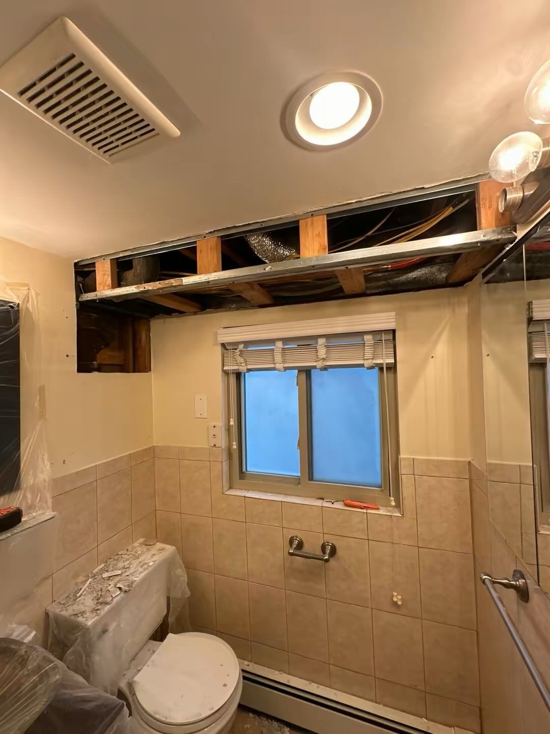 Bathroom with exposed ceiling framing, beige tiles, window, toilet, and shower area under renovation