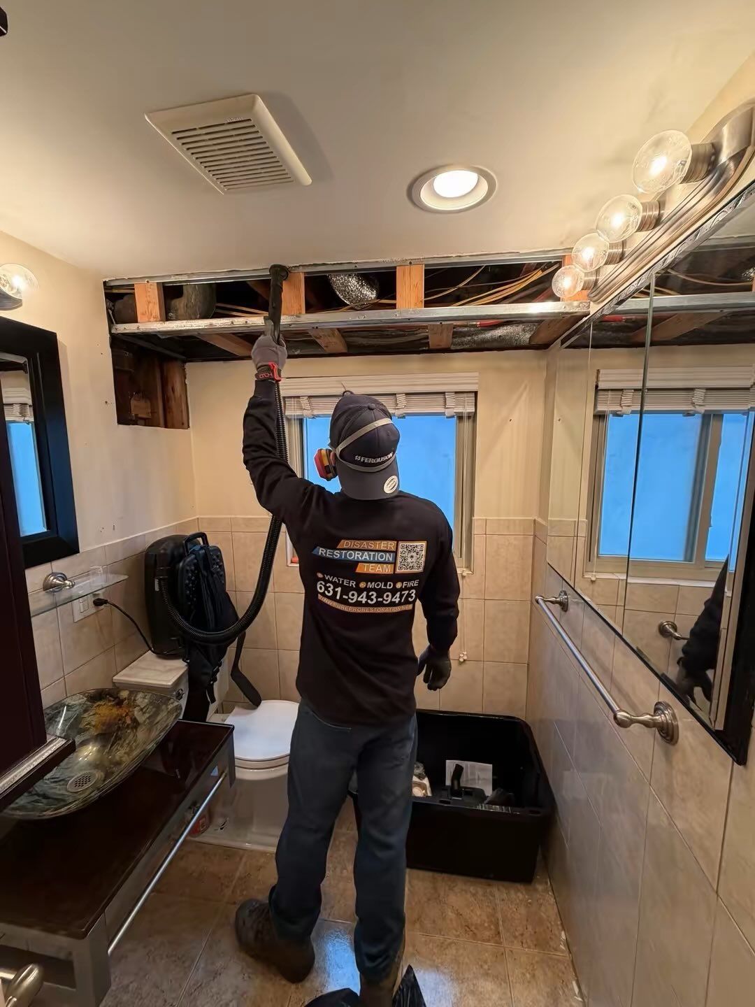 Person in a small bathroom removing ceiling panels during renovation, with exposed wiring and tools.