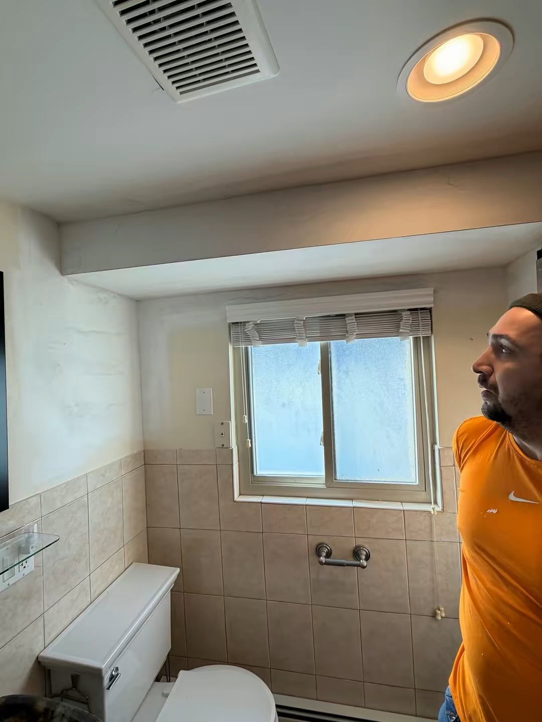 Small bathroom with a window, tub, and ceiling lights; a man in an orange shirt stands on the right.