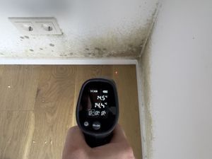 Handheld moisture meter reading a damp corner where a wood floor meets a white wall and ceiling stain