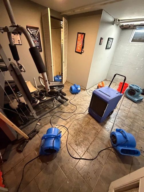 Basement room with workout gear, blue fans, cords, and a purple cube on tiled floor.