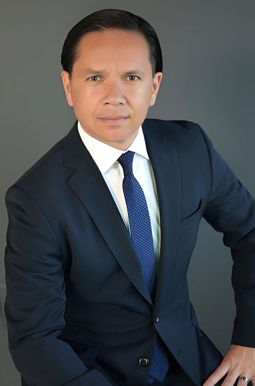 Studio portrait of a person in a navy suit and blue tie against a gray background