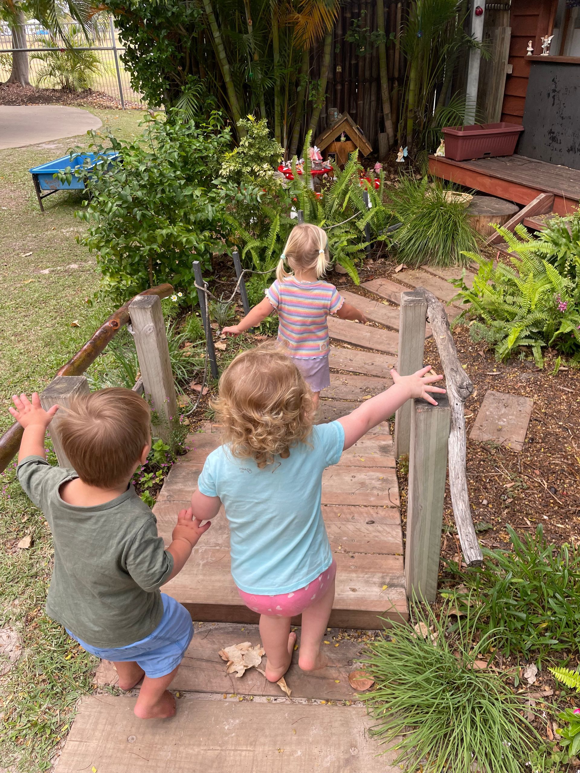 Three young children walking across a small wooden bridge in a garden, arms outstretched.