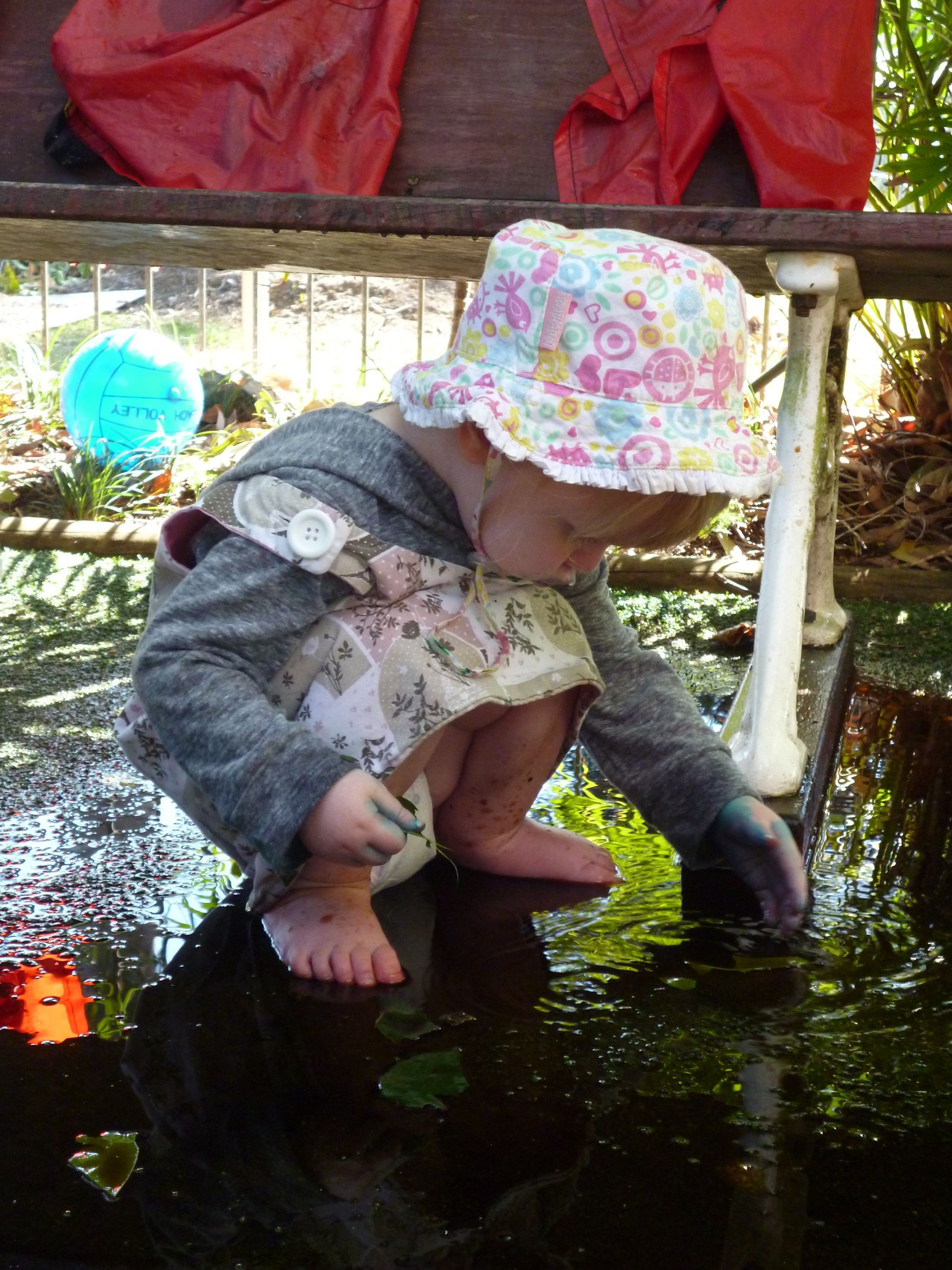 Child wearing a floral hat and gray sweater playing in dark water, feet and hands in the water.