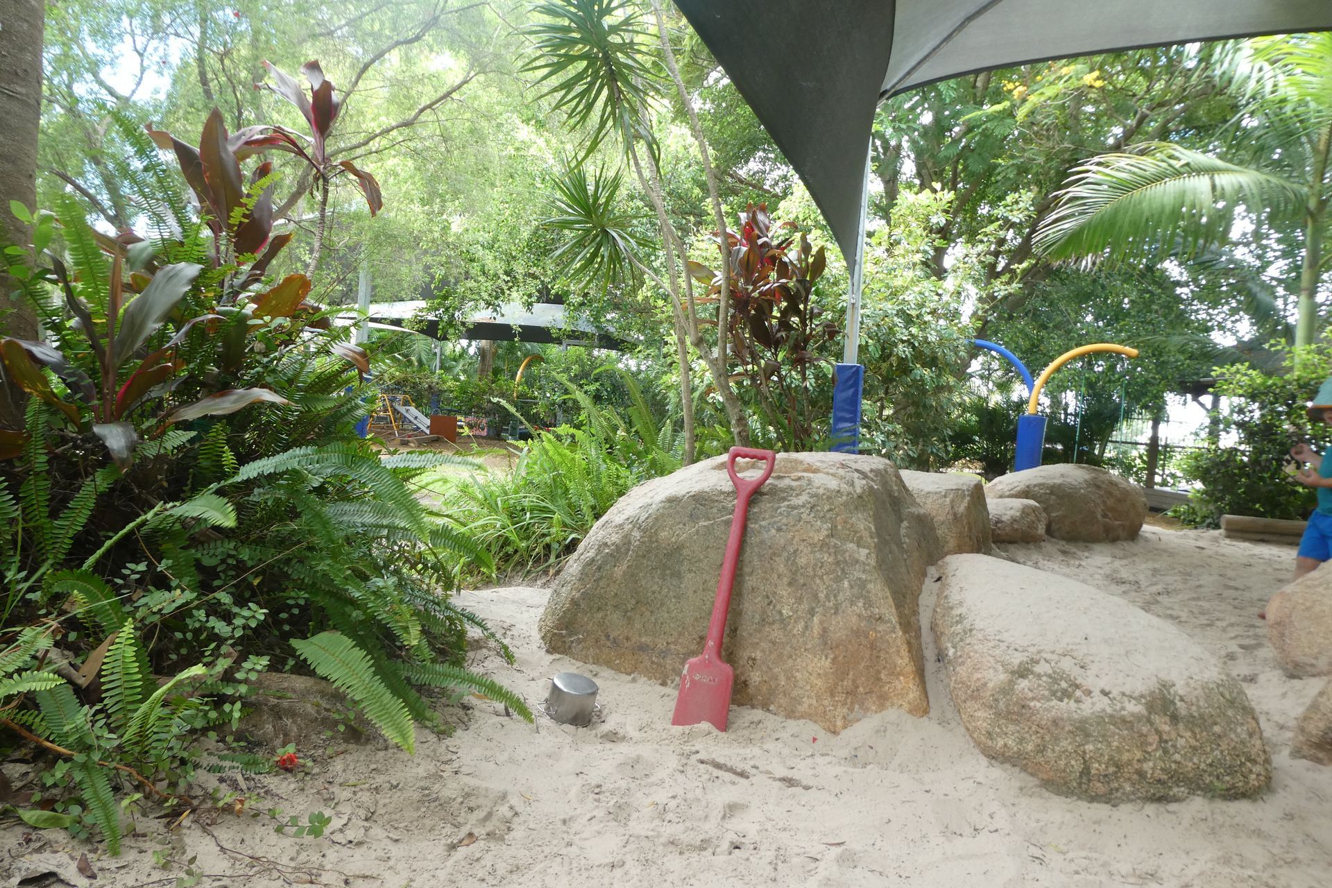 Sandpit with large rocks, shovel, and shade in a garden setting.