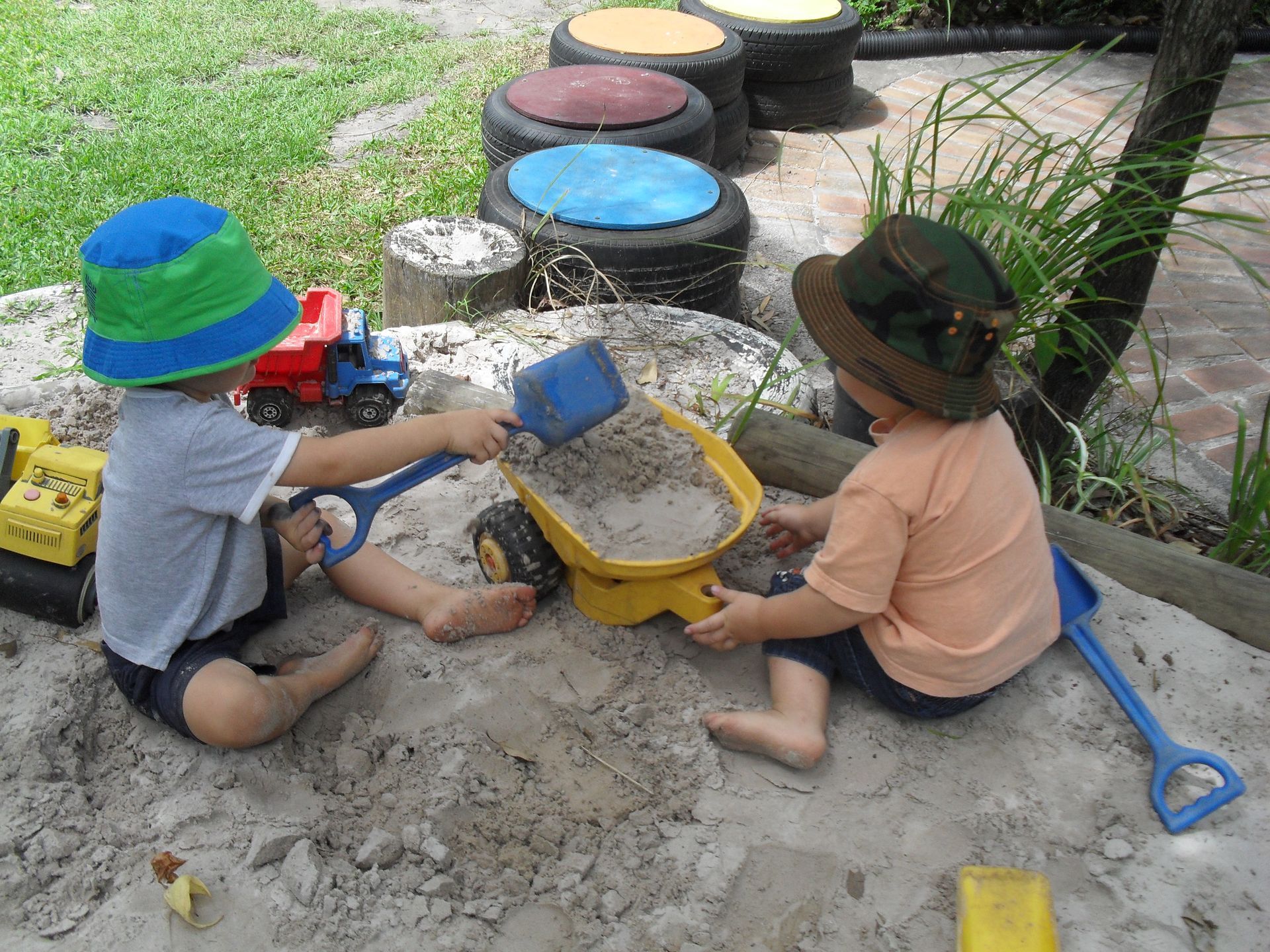 Two toddlers playing in a sandbox with trucks, shovels, and a wheelbarrow.