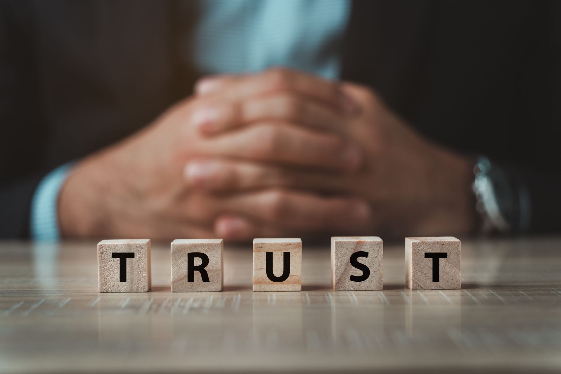 Hands clasped behind wooden blocks spelling “TRUST” on a table.