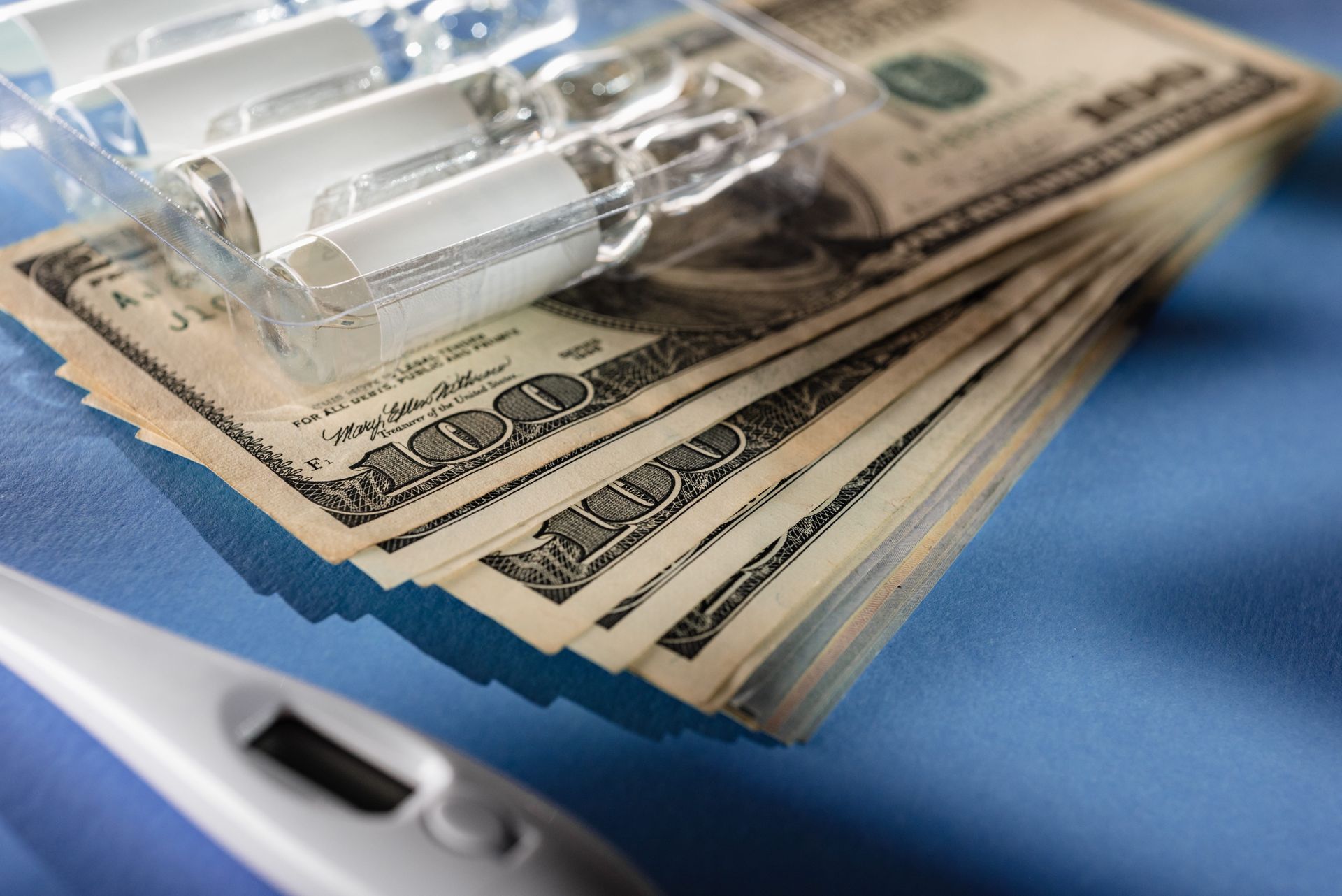 Clear glass medical ampoules in a plastic tray resting on top of a stack of US 100-dollar bills near a digital thermometer.