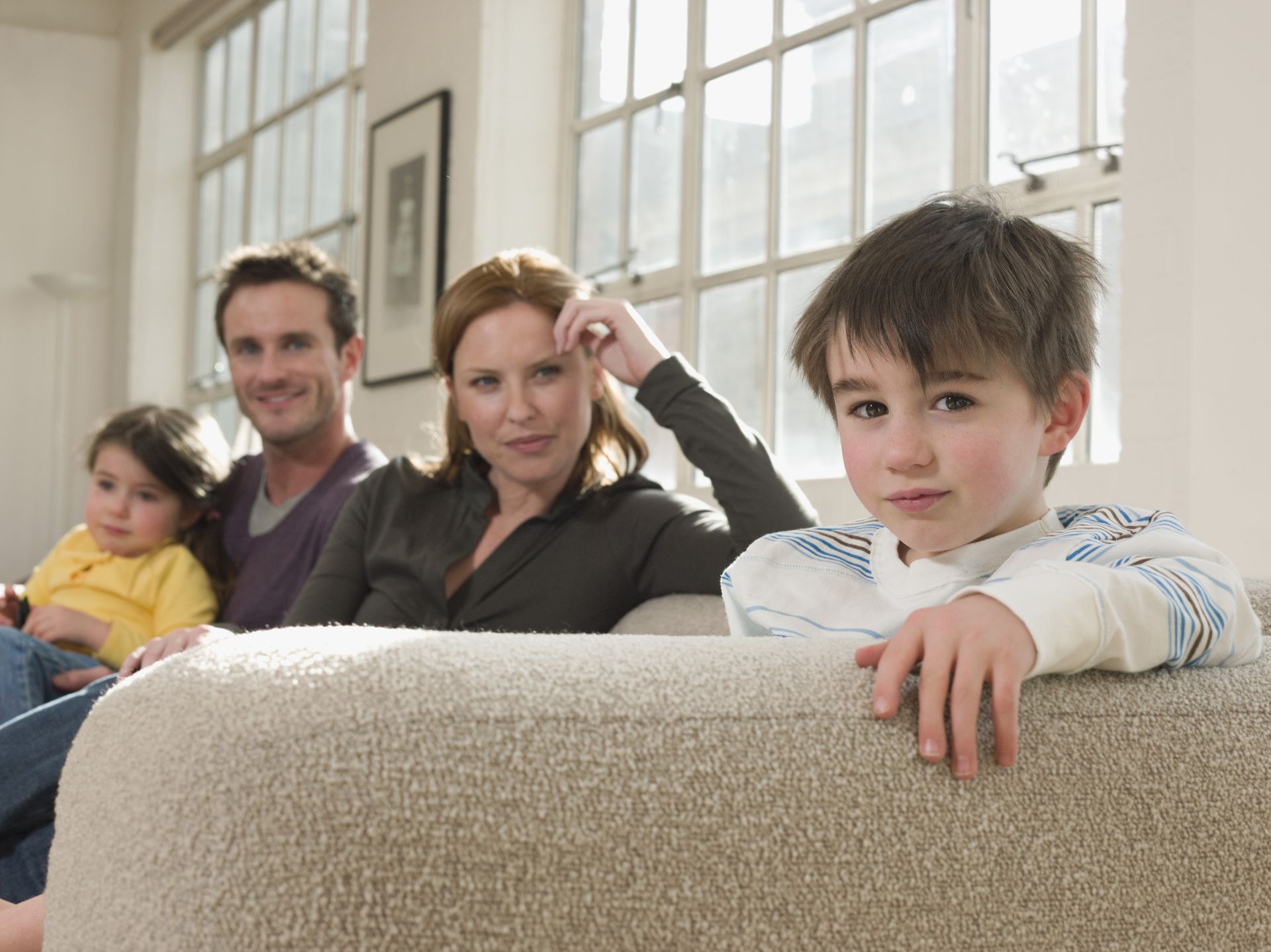 Family sitting on a couch. Boy in foreground, others in the background near windows.