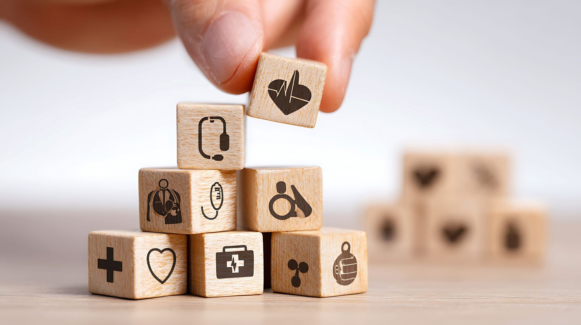 A hand places a wooden block with a heart and EKG icon on top of a stack of blocks featuring various medical-themed icons.