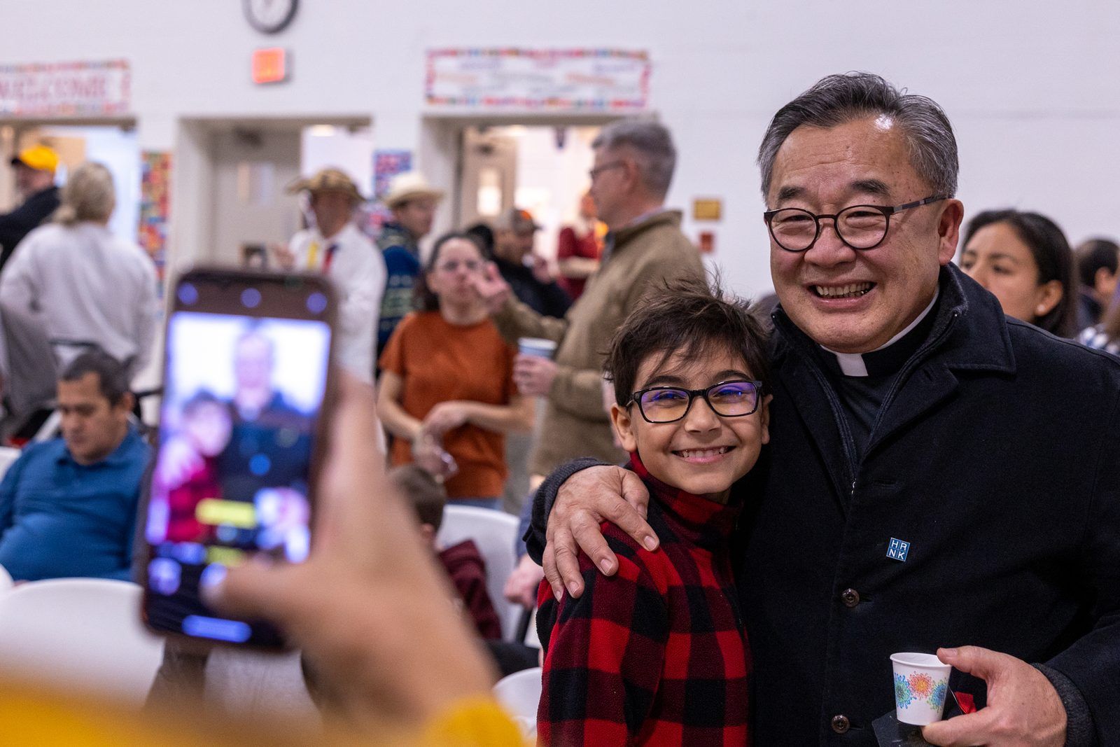 Smiling man and child posing together indoors while someone photographs them with a phone