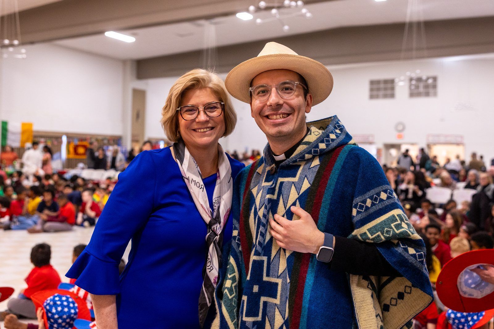 Two smiling people pose at an indoor event, one in a blue outfit and one in a straw hat and poncho.