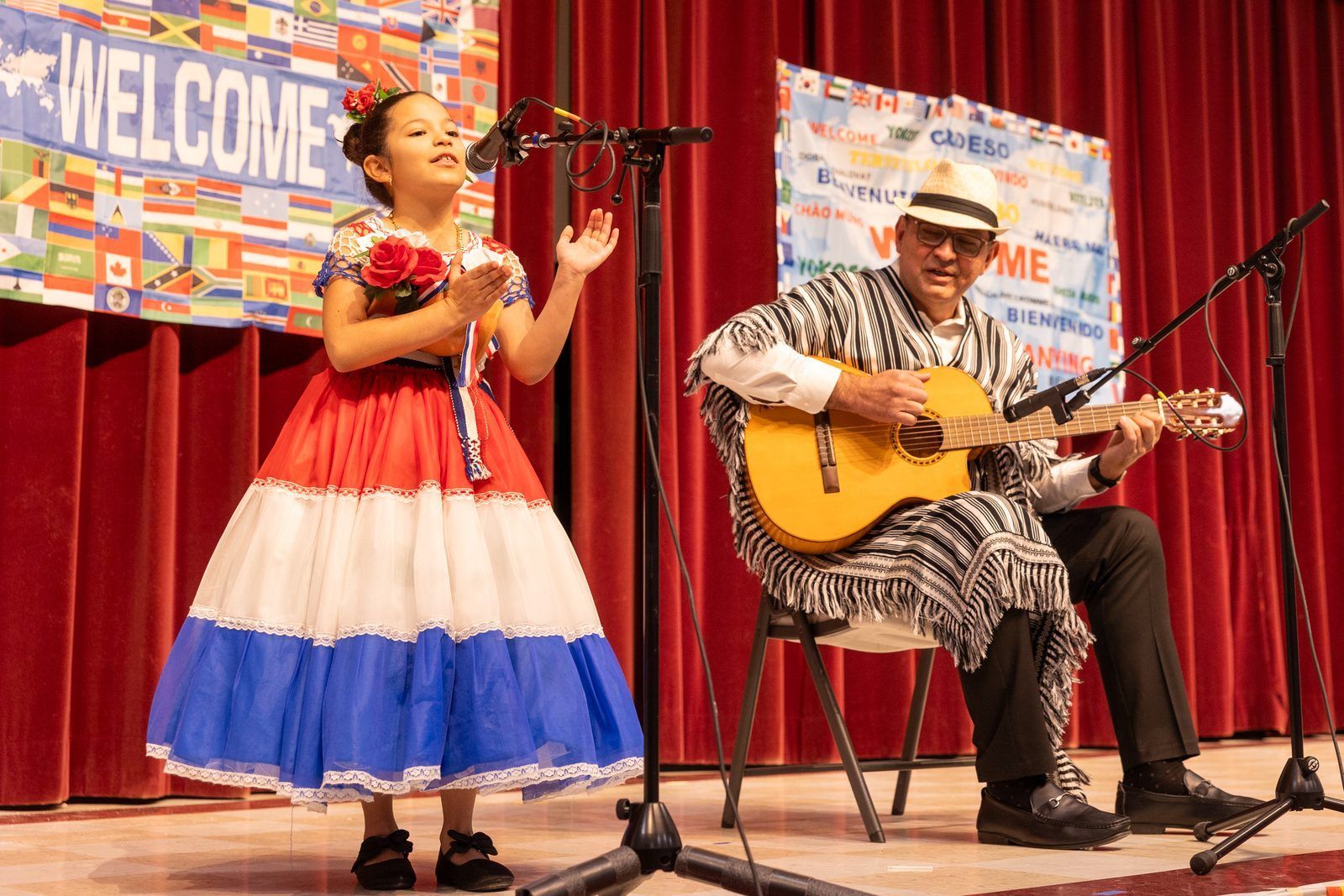 Two performers onstage: a woman singing in a red-white-blue dress and a man playing guitar in front of red curtains.